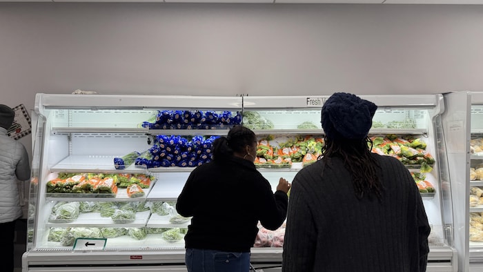 Deux employées de la banque alimentaire se préparent à donner des légumes aux usagers devant un réfrigérateur partiellement rempli. 