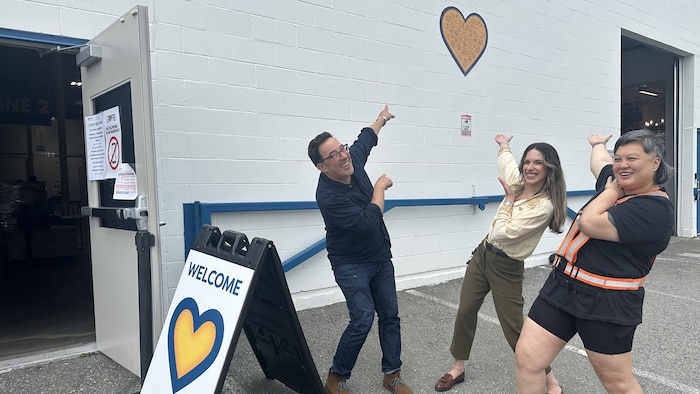 Trois employés de la Banque alimentaire du Grand Vancouver pointent de façon enjouée un coeur peint sur le mur du bâtiment.