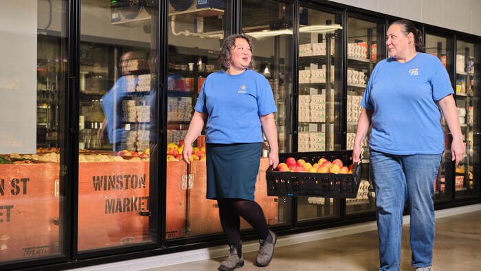 Deux employées transportent une caisse de pommes et marchent à côté d'énormes réfrigérateurs.