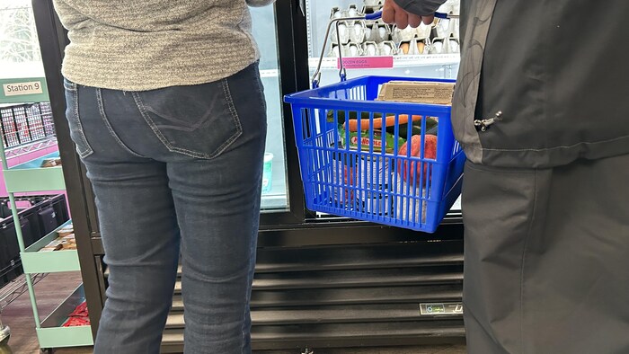 A man of whom only the middle of his body is visible holds a basket in front of a refrigerator. Another person is next to him and holds the fridge door open.