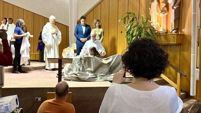 Une femme de dos prend une photo avec son cellulaire de sa fille, dans le choeur de l'église, assise dans une petite piscine, en train de se faire baptiser par un prêtre qui se trouve à genoux près de l'adolescente. 