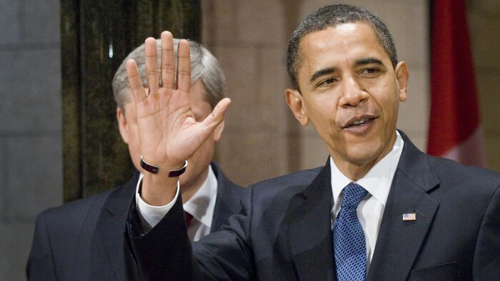 Barack Obama salue la foule et sa main cache partiellement le visage de Stephen Harper.