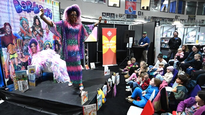 Barbada, une drag queen en robe scintillante anime une lecture de contes devant un groupe d'enfants attentifs.