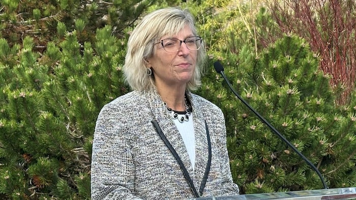 La ministre debout dans un parc, appuyée sur un lutrin, devant des conifères.