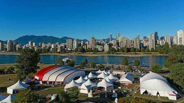 Plusieurs tentes de tailles différentes au bord de l'eau avec une ville et des montagnes en arrière-plan.