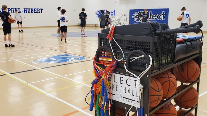 Des adolescents jouent au basketball en arrière-plan d'un bac de matériel sportif à l'école Paul-Hubert de Rimouski.