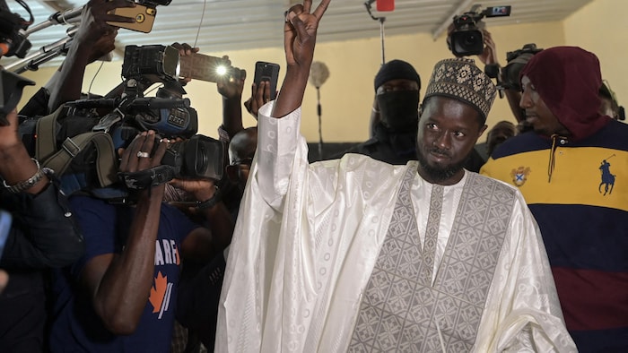 Bassirou Diomaye Faye fait un signe de paix avec sa main; il est entouré de caméras.