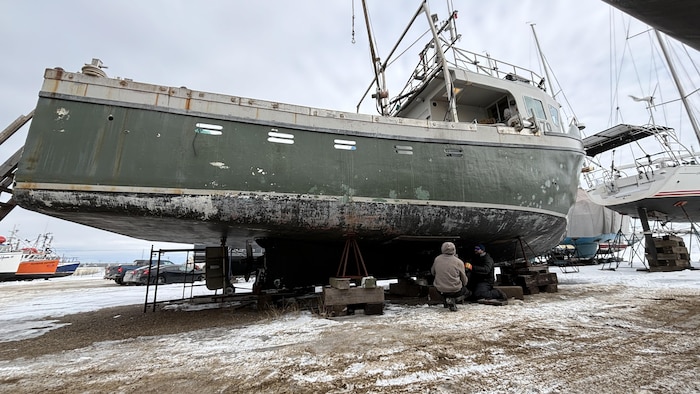 Deux hommes sont agenouillés près de la coque d'un bateau de pêche installé sur des tréteaux. 