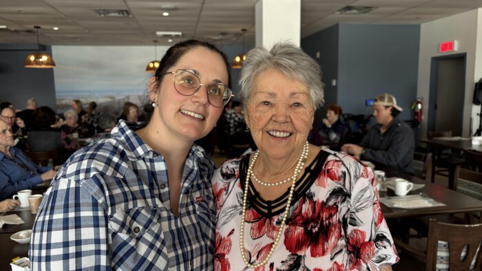 Deux femmes sourient à la caméra dans une salle à manger pleine de gens attablés.
