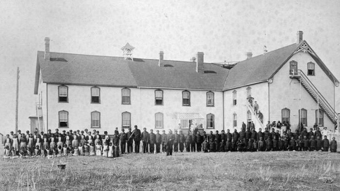 Des élèves debout devant le pensionnat autochtone Battleford Industrial School en 1895 qui comprend deux étages.