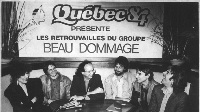 Les membres du groupe sont assis autour d'une table, souriants et d&eacute;tendus, sous une grande affiche &laquo; Qu&eacute;bec 84 pr&eacute;sente Les retrouvailles du groupe Beau Dommage &raquo;.