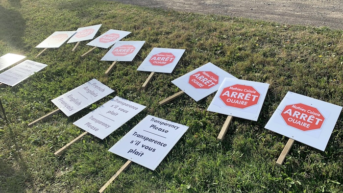 De petites affiches carrées sont posées à terre sur la pelouse. Certaines disent « Transparence s'il vous plaît » et les autres ont un signe d'arrêt-stop et les mots « Mathieu Caissie arrêt ouaire ».