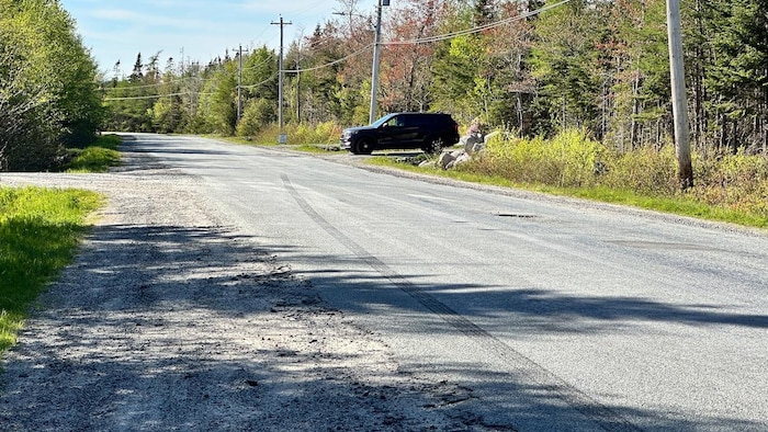 Une fourgonnette noire est stationnée devant un boisé, perpendiculairement à une route étroite.