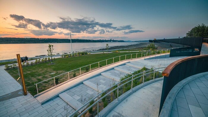 Photographie en grand-angle d'une promenade publique au crépuscule. Au premier plan, des escaliers en béton clair avec rampes métalliques serpentent vers une pelouse verdoyante. Un sentier accessible longe le fleuve calme. Le ciel affiche des teintes orangées et bleues avec quelques nuages. À l'horizon, on devine la silhouette d'une ville et des collines sous une lumière douce de fin de journée.