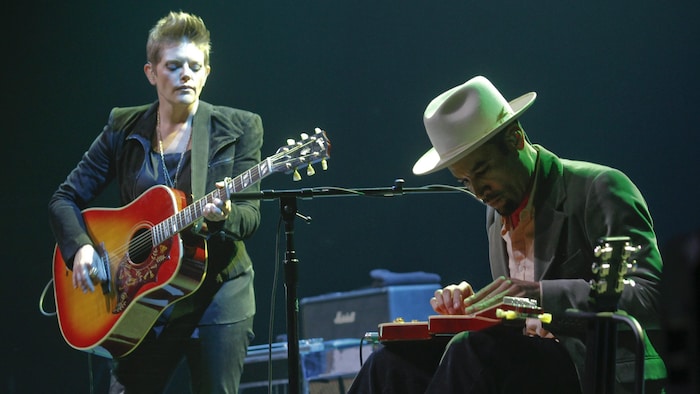 Coiffé d'un élégant chapeau blanc, Ben Harper joue de la slide-guitare, assis et penché sur son instrument. Natalie Maines est debout à sa gauche et joue de la guitare.