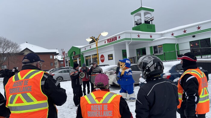 Une scène hivernale montre un groupe de personnes, incluant des agents de surveillance en dossards orange et une mascotte bleue, devant un bâtiment Texaco. Le toit est orné d'une réplique de la DeLorean du film « Retour vers le futur ».