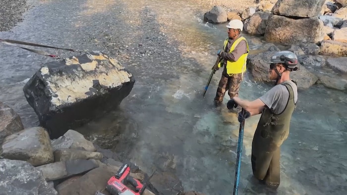 Deux hommes regardent un rocher être tiré par une corde dans une rivière.