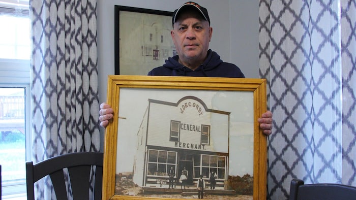 Un homme coiffé d'une casuquette tient une photo antique d'un magasin appelé J. Decorby General Merchant.