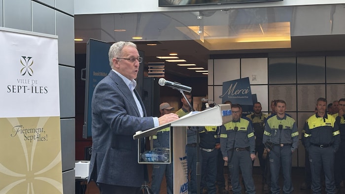 Benoit Méthot parle avec conviction dans un microphone à un lutrin. En arrière-plan, plusieurs travailleurs d'Aluminerie Alouette en uniformes de travail écoutent attentivement.