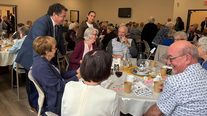 Une grande salle pleine de personnes âgées pleine de tables. Un homme et une femme debouts discutent avec des personnes assises.