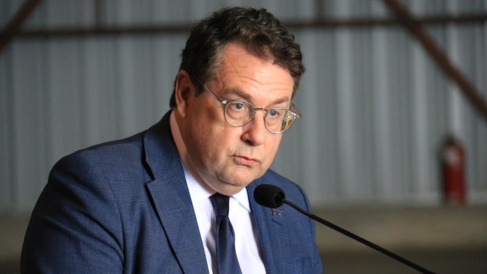 Bernard Drainville, avec des lunettes et un costume, parle sérieusement derrière un micro.