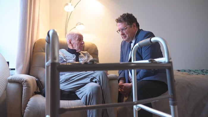 Un homme en veston est assis au pied d'un lit, à côté d'un monsieur âgé assis dans un fauteuil. En avant-plan, on aperçoit la marchette de l'homme.