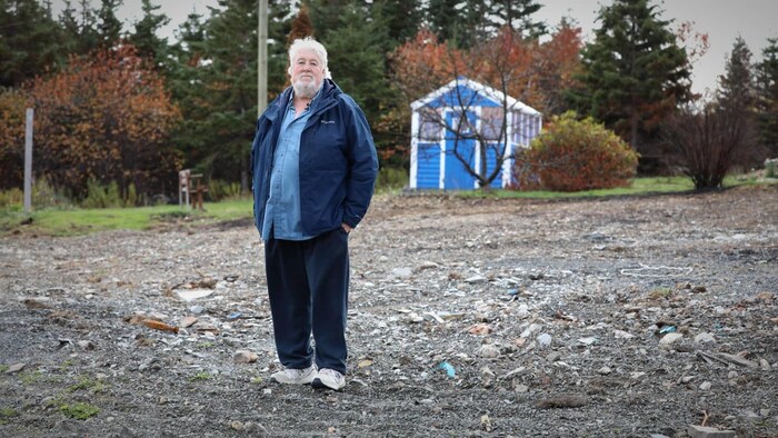 Un homme aux cheveux blancs et à la barbe blanche debout sur un terrain où une maison a brûlé. Il y a un cabanon intact derrière.