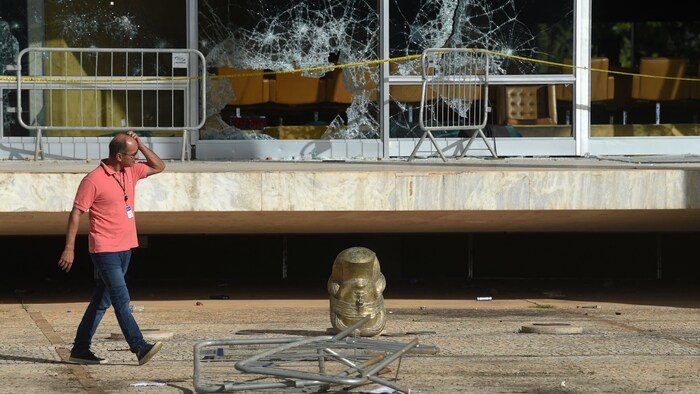Un homme marche près de la tête coupée d'une statue renversée sur le sol. Des fenêtres sont brisées derrière lui.