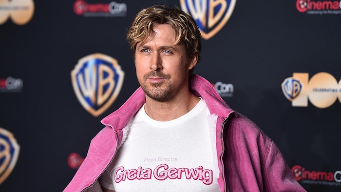 Ryan Gosling pose sur le tapis rouge, avec un manteau rose qu'il tient ouvert. Sur son t-shirt, on peut lire en anglais «De la réalisatrice Greta Gerwig». 