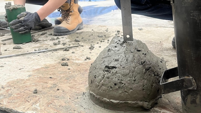 Une personne mesure à l'aide d'une règle l'affaissement d'un amas de béton en forme de cône sur le sol.