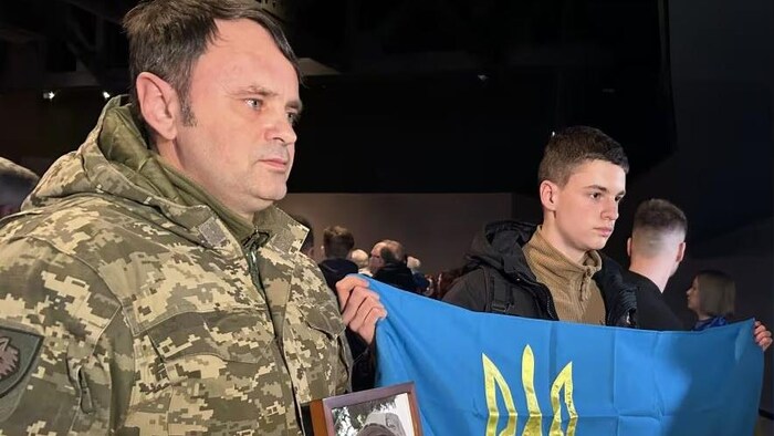 Betro Lukin tient une photo de son fils aîné alors qu'un autre de ses fils garde le drapeau de l'Ukraine à côté de lui, le 23 février 2025 à Winnipeg.