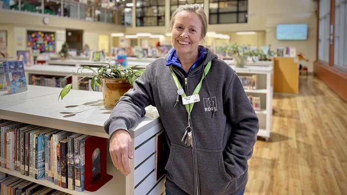 Une femme se tient debout à côté d'une étagère de livres et sourit.