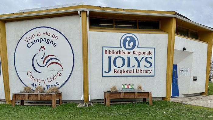 L'extérieur du bâtiment de la Bibliothèque régionale Jolys. Une pancarte indique les paroles « Vive la vie en campagne ».