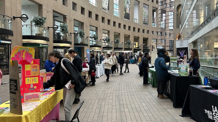 Dans la bibliotheque centrale de Vancouver, des dizines de tables se trouveux d'un côté et de l'autre d'un couloir. Des gens se trouvent derrière les tables, et d'autres passent devant.