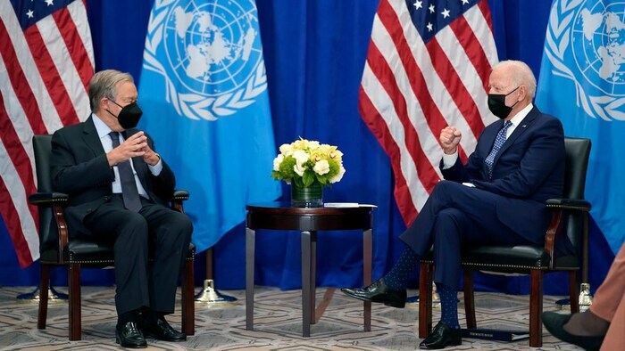 U.S. President Joe Biden met with UN Secretary General Antonio Guterres. They are both sitting on chairs, with a small table between them. United Nations and American flags in background.