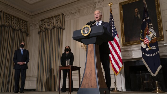 Joe Biden parle aux reporters devant un drapeau américain et un portrait d'Abraham Lincoln, sous le regard de John Kerry et de Kamala Harris.