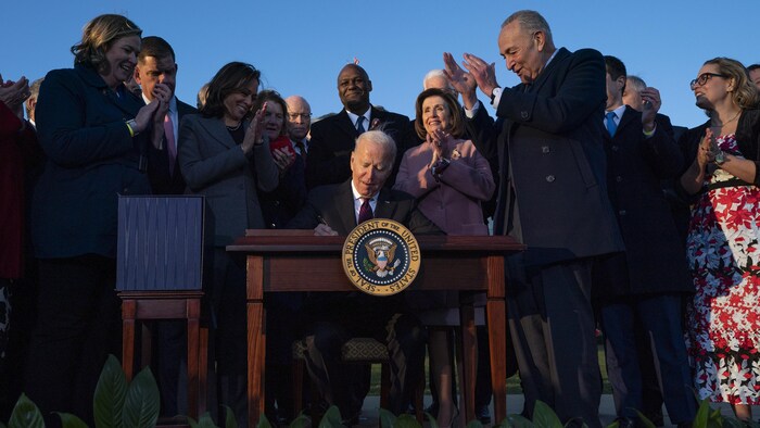 Joe Biden, assis, signe le texte de loi, applaudi par les élus, debout, autour de lui, dont Kamala Harris, Nancy Pelosi et Chuck Schumer, souriants.