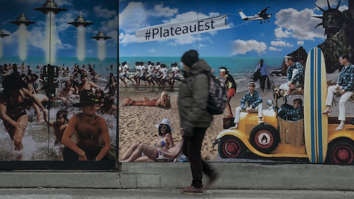 Un homme masqué passe devant une murale représentant pour moitié une attaque extraterrestre sur une plage, pour l'autre des gens qui bronzent sur une plage.