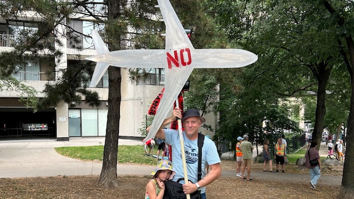 Steve Richards tient bien haut la silhouette d’un avion de ligne sur lequel on pourrait lire « non »  en lettre rouge avec qu’une fillette s’accroche à sa jambe. 