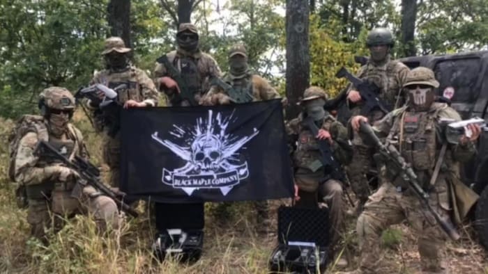 Sept hommes armés et masqués autour d'un drapeau avec l'insigne de la Black Maple Company.