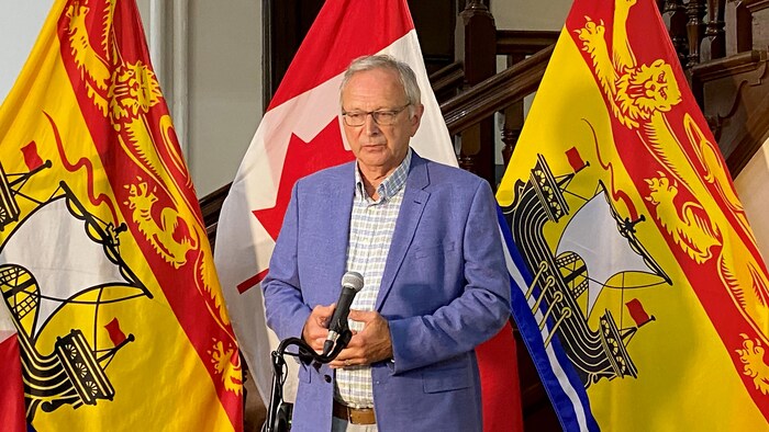 Blaine Higgs debout devant un drapeau du Canada et deux drapeaux du Nouveau-Brunswick.