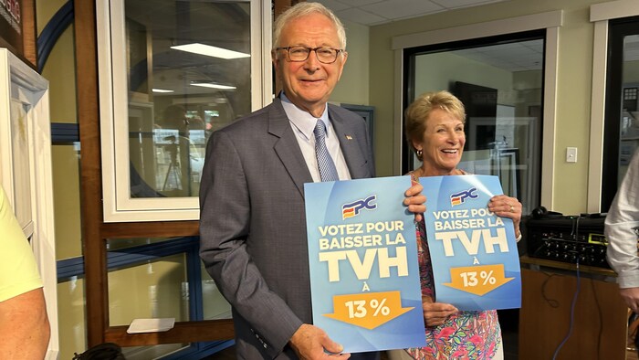 Un homme et une femme tenant chacun une affiche indiquant : « Votez pour baisser la TVH à 13 % ».