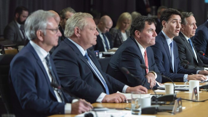 Des messieurs en complet assis côte à côte à une même table, photographiés de profil, des documents et des micros devant eux. Justin Trudeau est en train de parler.