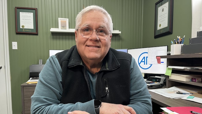 Blair Aitken est assis à son bureau. Il regarde la caméra.