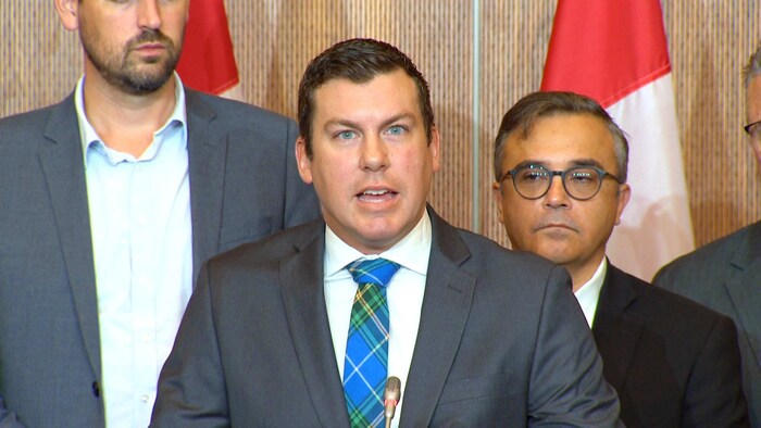 Kody Blois parle flanqué de deux individus, dont l'un est si grand de taille qu'il a la tête coupée par la bordure de la photo.