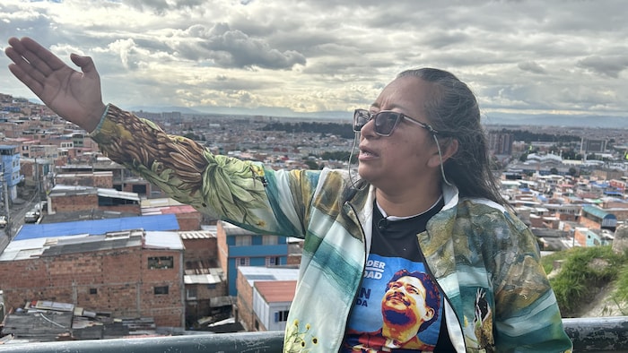Une femme pointe vers la gauche en parlant devant un appareil photo. En arrière-plan, la ville de Bogota.