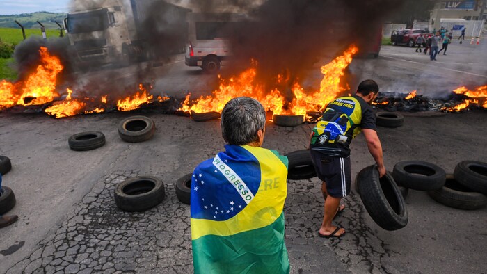 Un hombre quema neumáticos en la calle mientras otro hombre envuelto en una bandera brasileña mira.