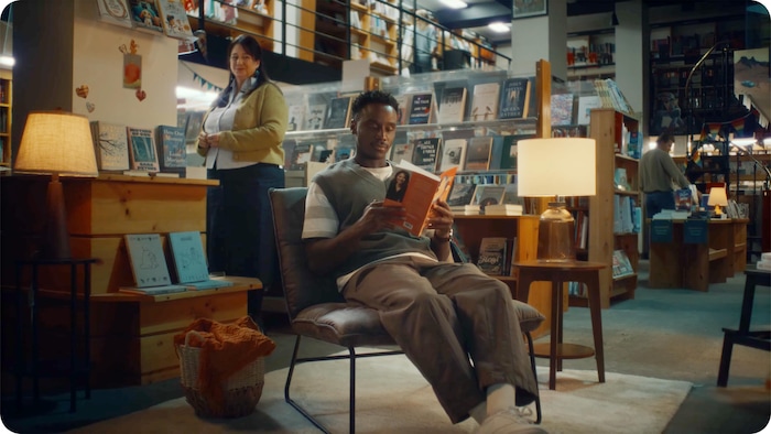 Un homme lit un ouvrage assis dans un fauteuil au cœur d'une librairie chaleureuse éclairée par des lampes d'appoint.