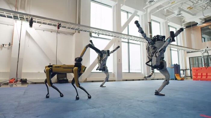 Capture d'écran d'une vidéo montrant le robot-chien Spot et deux robots bipèdes Atlas de Boston Dynamics se déhancher dans un gymnase.