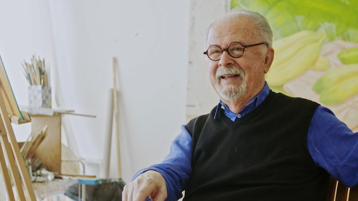 Un homme aux cheveux gris qui porte des lunettes rondes, assis dans un studio de peinture.
